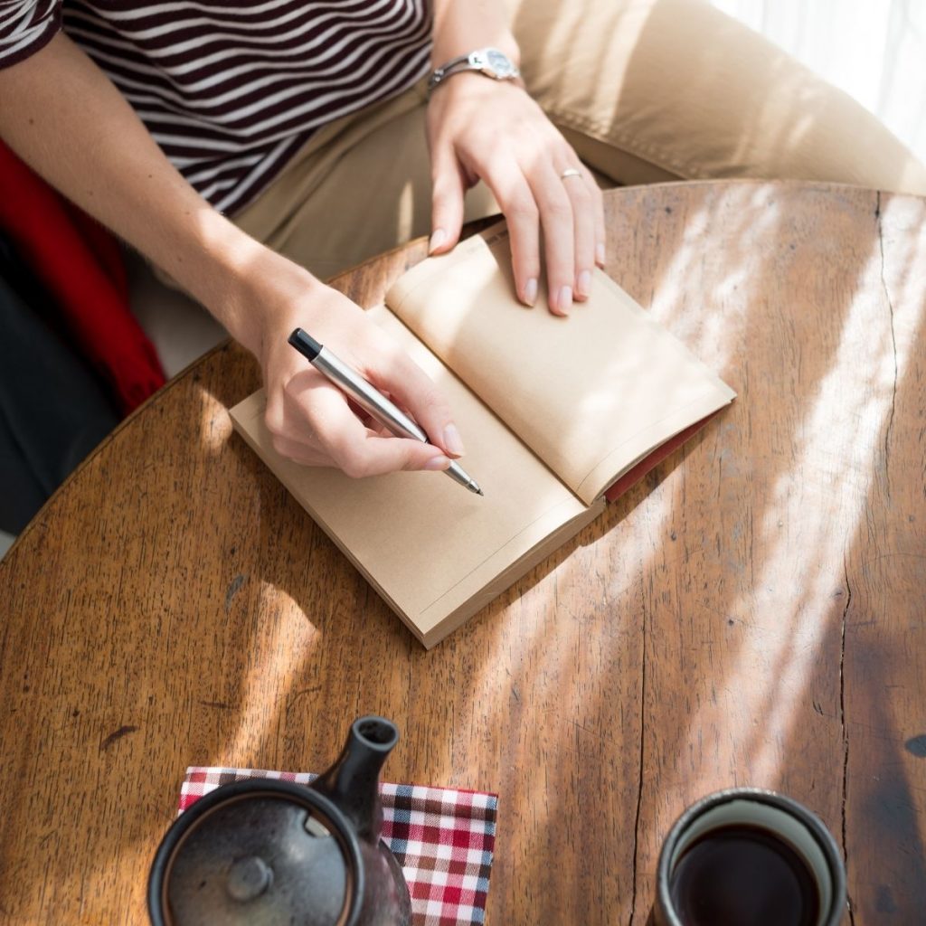 person in brown jeans and striped shirt journaling with tea pot on table